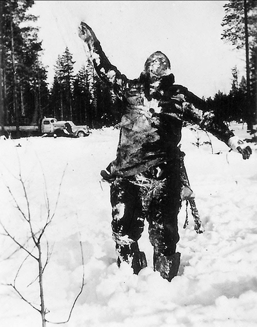 Замёрзшее тело советского военнослужащего стоит в снегу у лесной дороги; позади виден грузовик