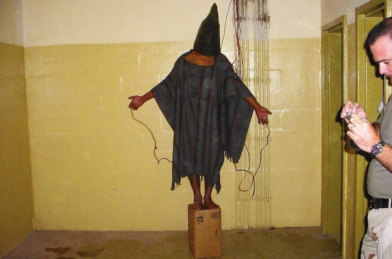 In Sabrina Harman’s photograph, Ivan Frederick stands beside the prisoner, looking at a digital camera in his hands. The CID caption dates the frame to about 11:04 p.m. on 4 November 2003.