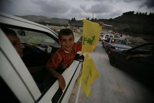 A boy holding a yellow Hezbollah flag leans out of a car window in a convoy.