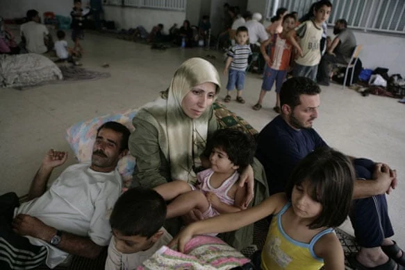 Adults and children sit on the floor beside their belongings; other people lie on mattresses behind them.