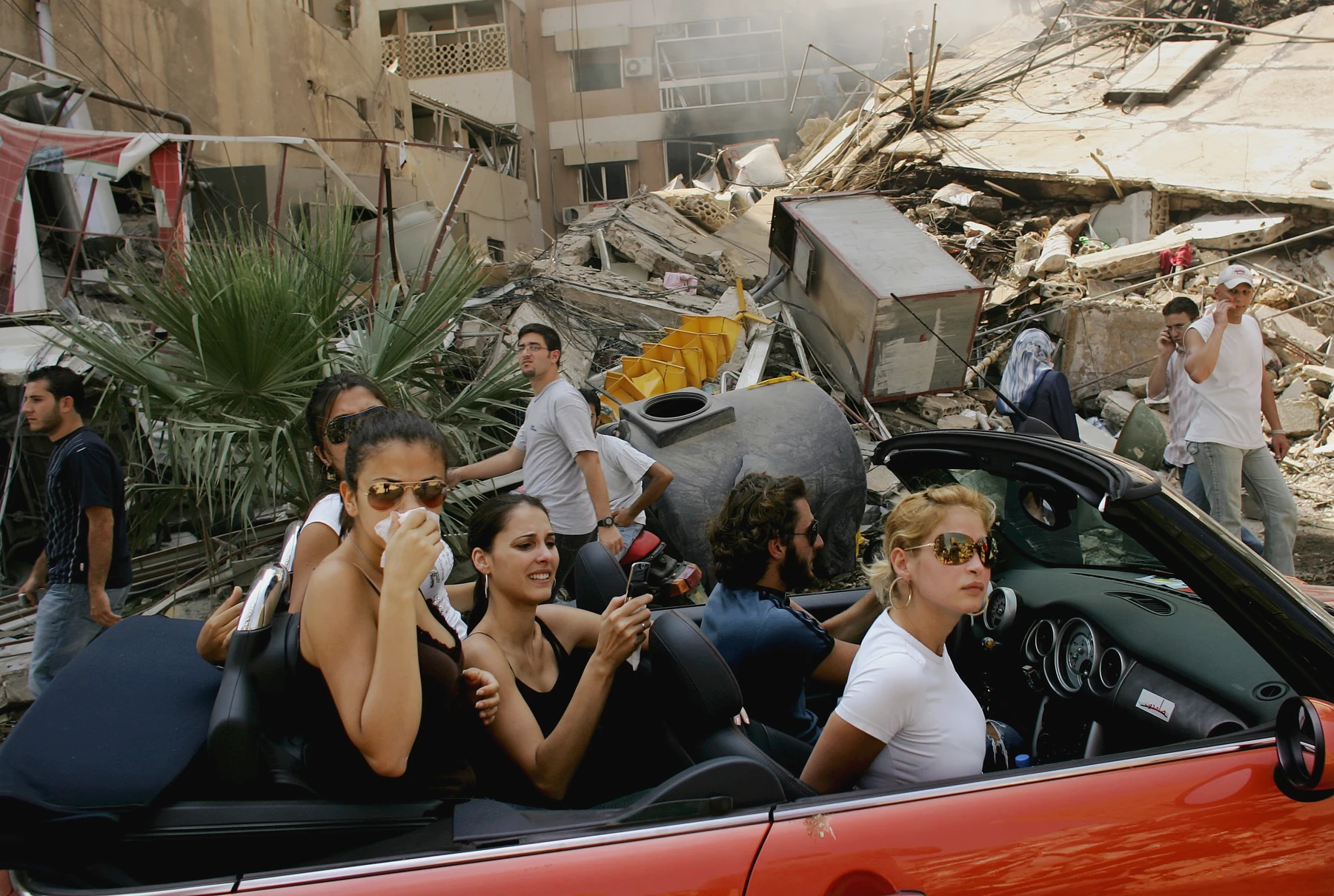 Five young people in a red convertible travel along a street amid ruined buildings.