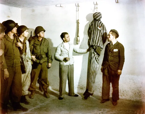 Two former prisoners stand before a group of soldiers beside a suspended dummy in striped clothing; one gestures towards it