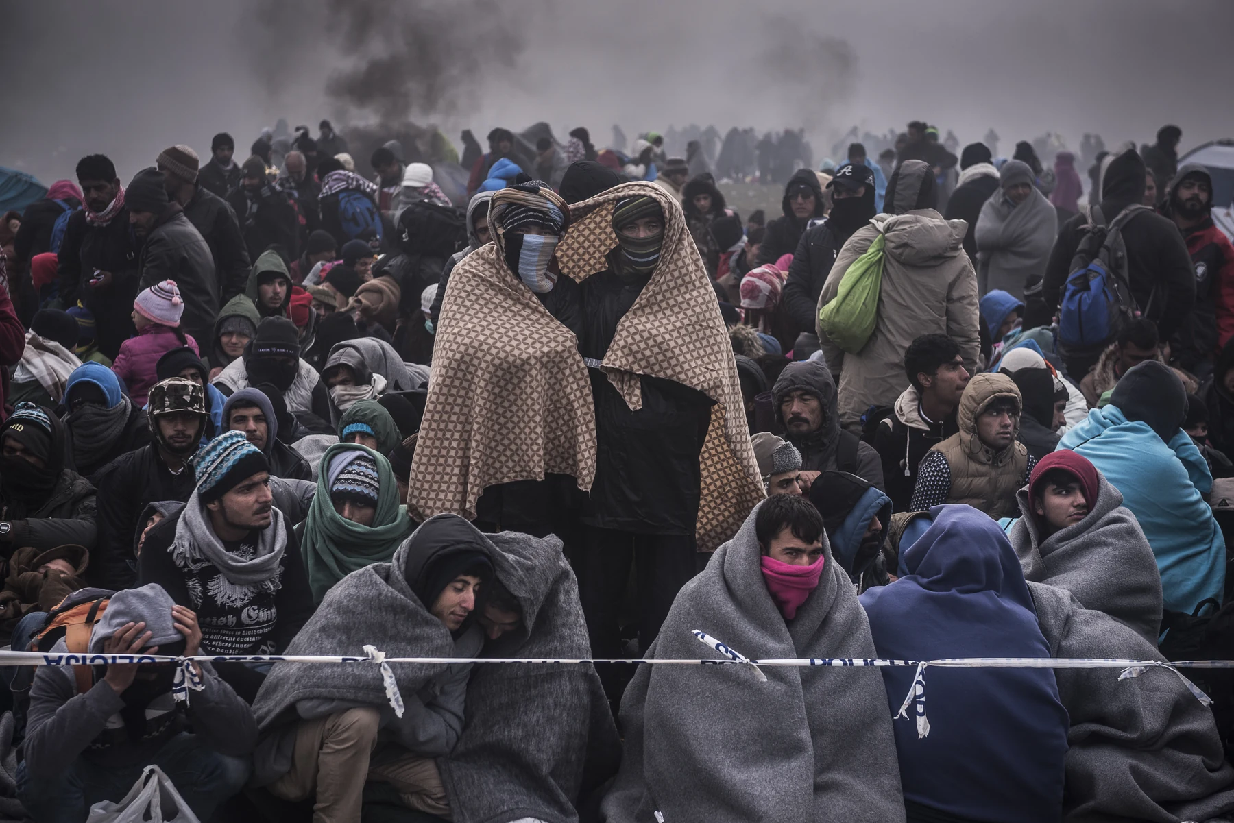 Adults and children, many wrapped in blankets, wait behind police tape.
