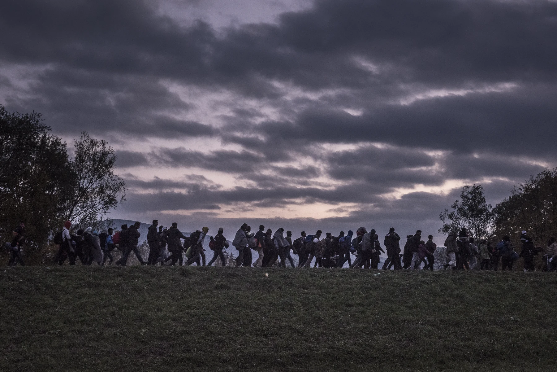 Silhouettes of people walk along an embankment beneath a dark, cloudy sky.