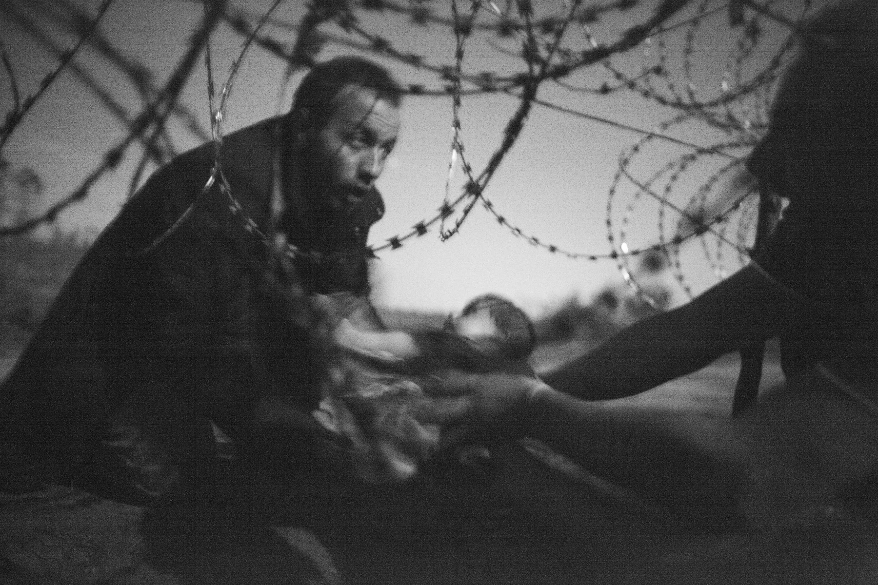 At night, adults pass a small baby through a gap in a wire fence.