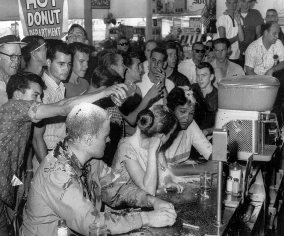 Three sit-in participants at the Woolworth’s counter are surrounded by a crowd; a man pours liquid