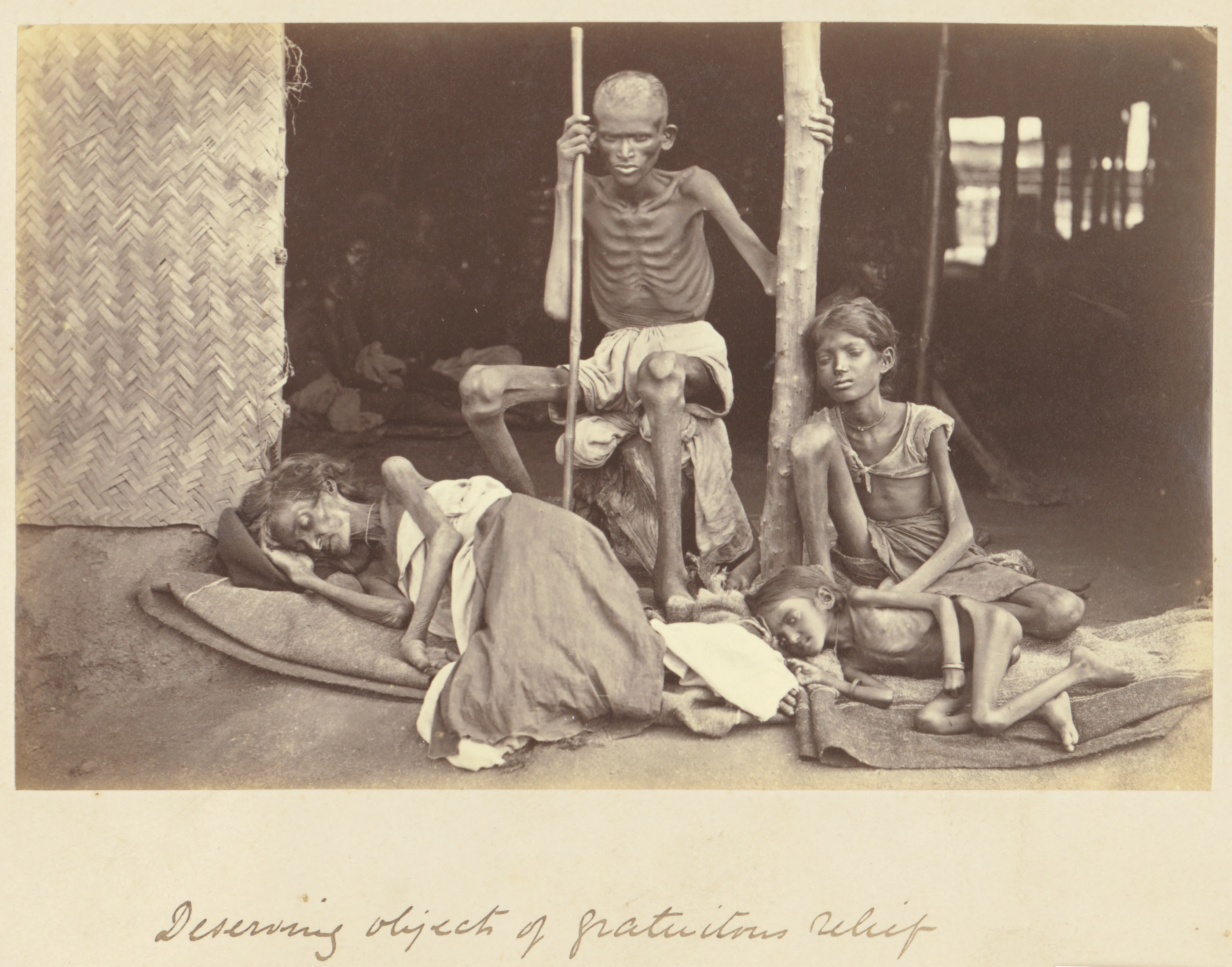 Four emaciated people at the entrance to a building: two sit and two lie on mats. The Getty Museum describes them as a family at a famine-relief camp.