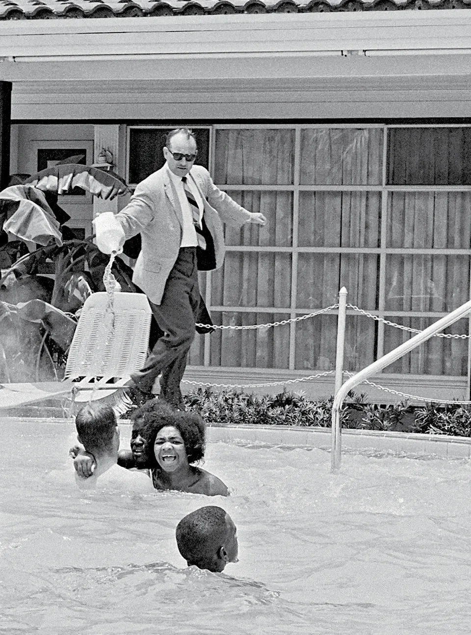 A suited man pours liquid from a container beside a pool with several protesters in the water