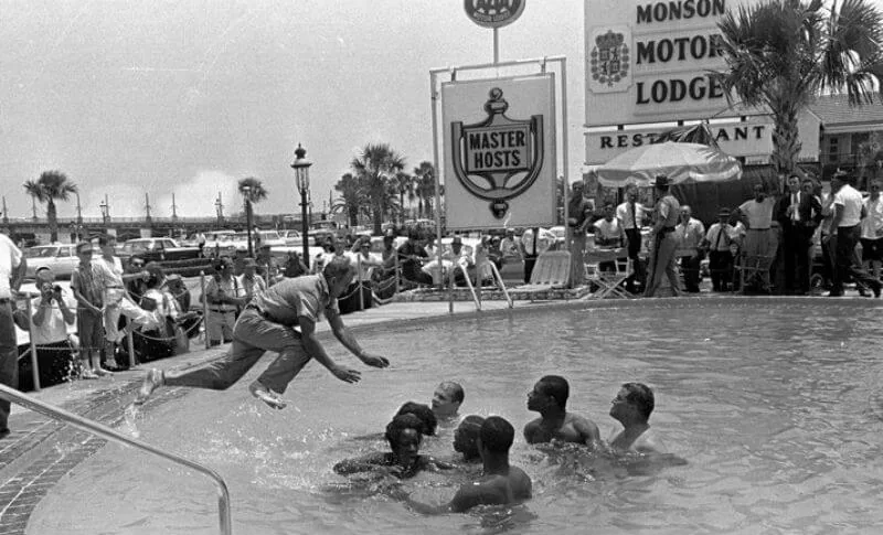 A clothed man jumps into a pool towards a group; a Monson Motor Lodge sign stands behind them