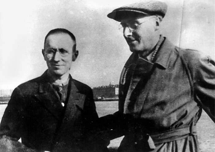 Two men stand together outdoors; the man on the right wears glasses, a cap and a belted coat.