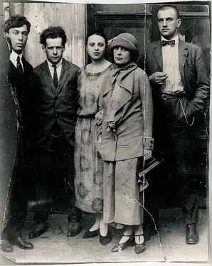 Five people by doors on an old photographic print with irregular edges and creases.
