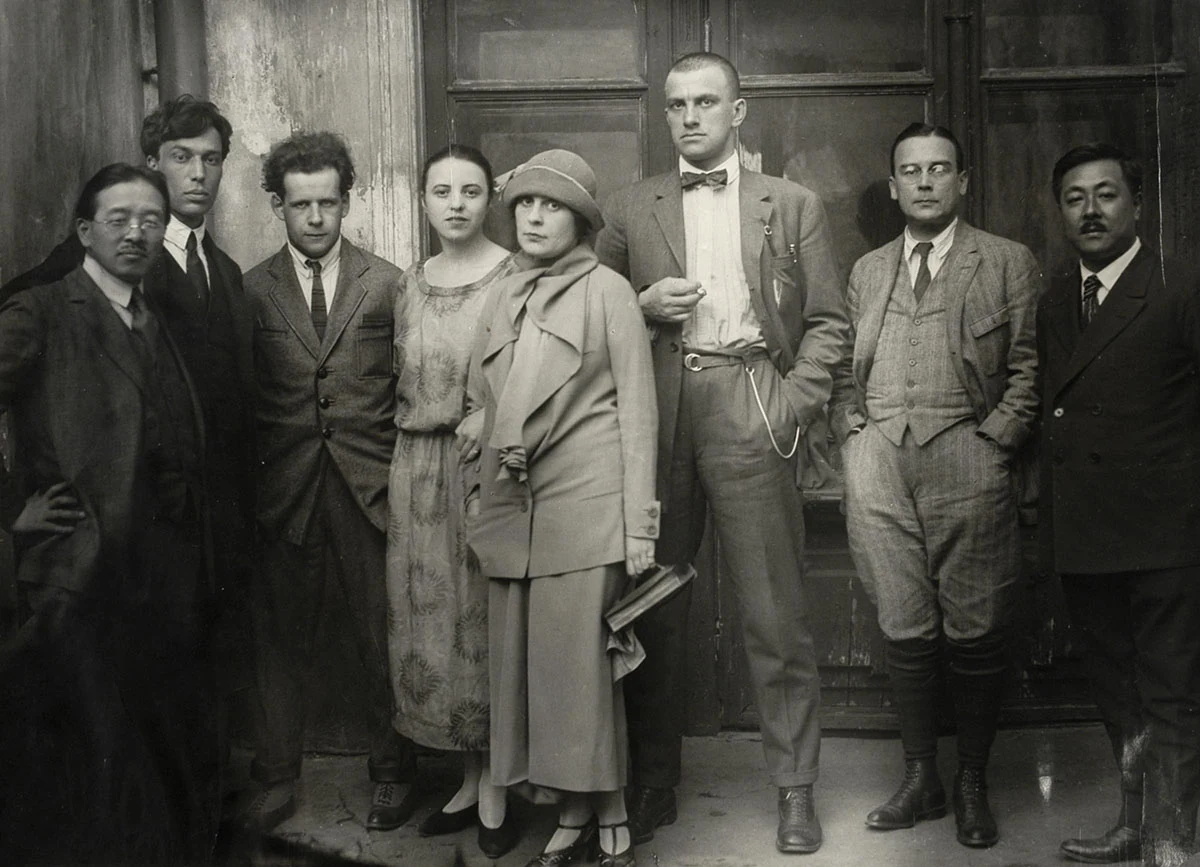 Eight people stand by wooden doors. Lilya Brik, wearing a hat and coat, stands in front of Mayakovsky.