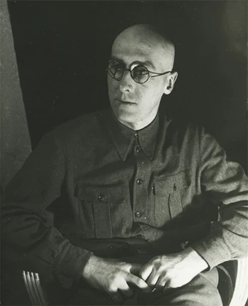 A man in round glasses and a dark buttoned shirt sits with his hands folded.