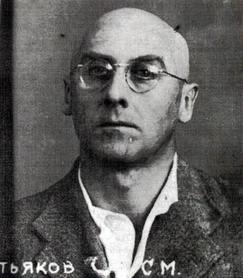 A head-and-shoulders portrait of a man in glasses, with part of a handwritten surname visible below.
