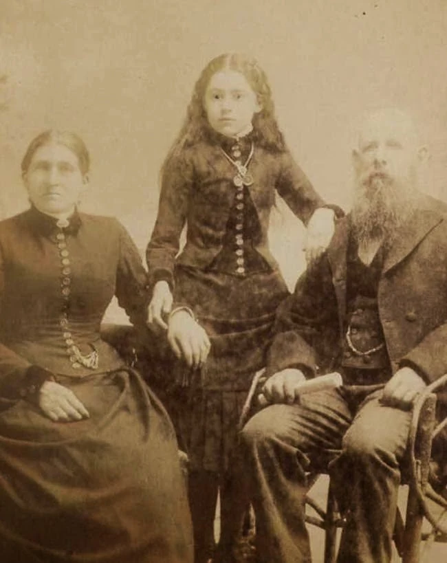 A girl stands between a seated man and woman, touching one adult’s hand and the other’s shoulder. Online lists call this a post-mortem portrait; no archival evidence confirms that claim or identifies the sitters.