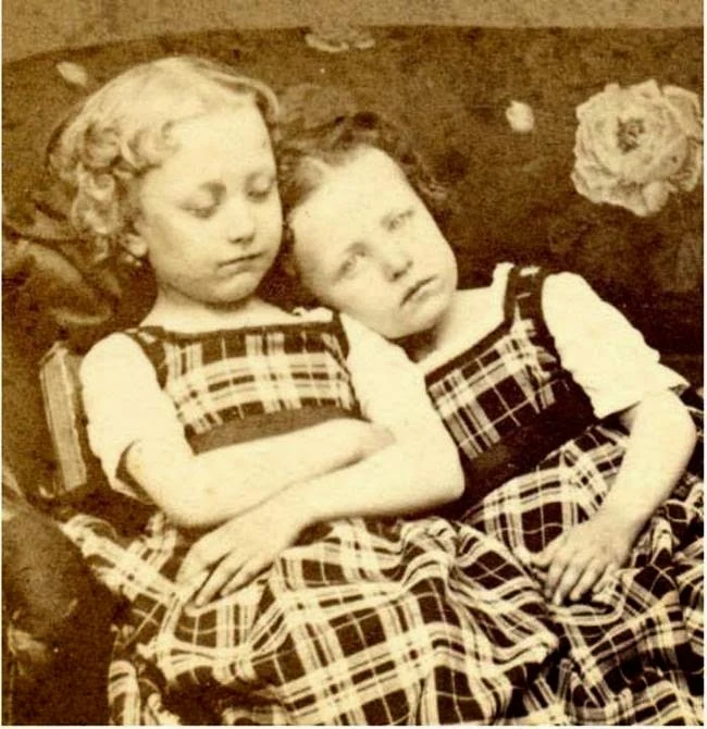Two girls in matching checked dresses sit leaning together. This portrait appears in post-mortem collections, but captions differ and no document establishes that either girl had died.