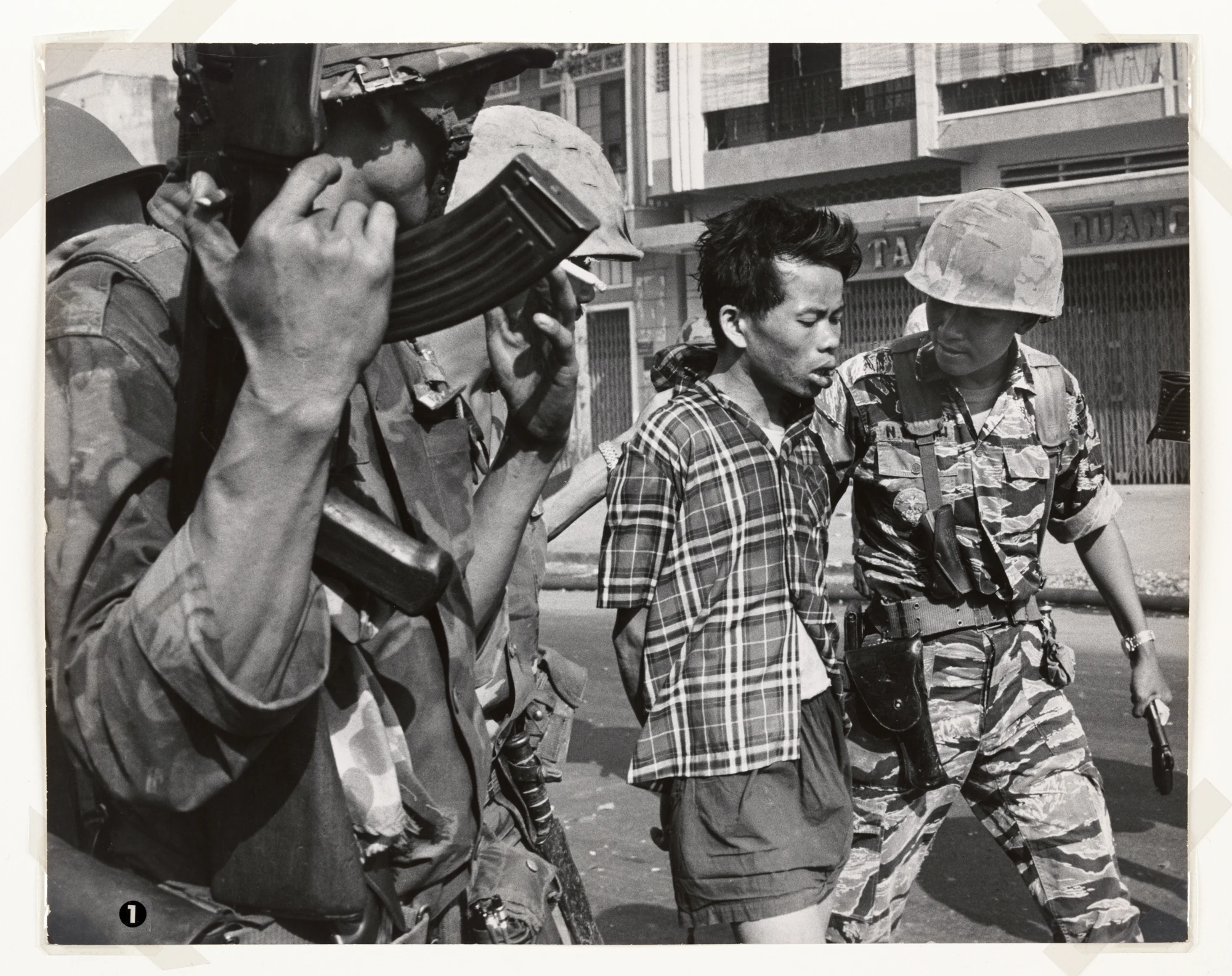 South Vietnamese soldiers escort Nguyễn Văn Lém. He wears a checked shirt and his hands are bound behind his back. Eddie Adams / Associated Press.