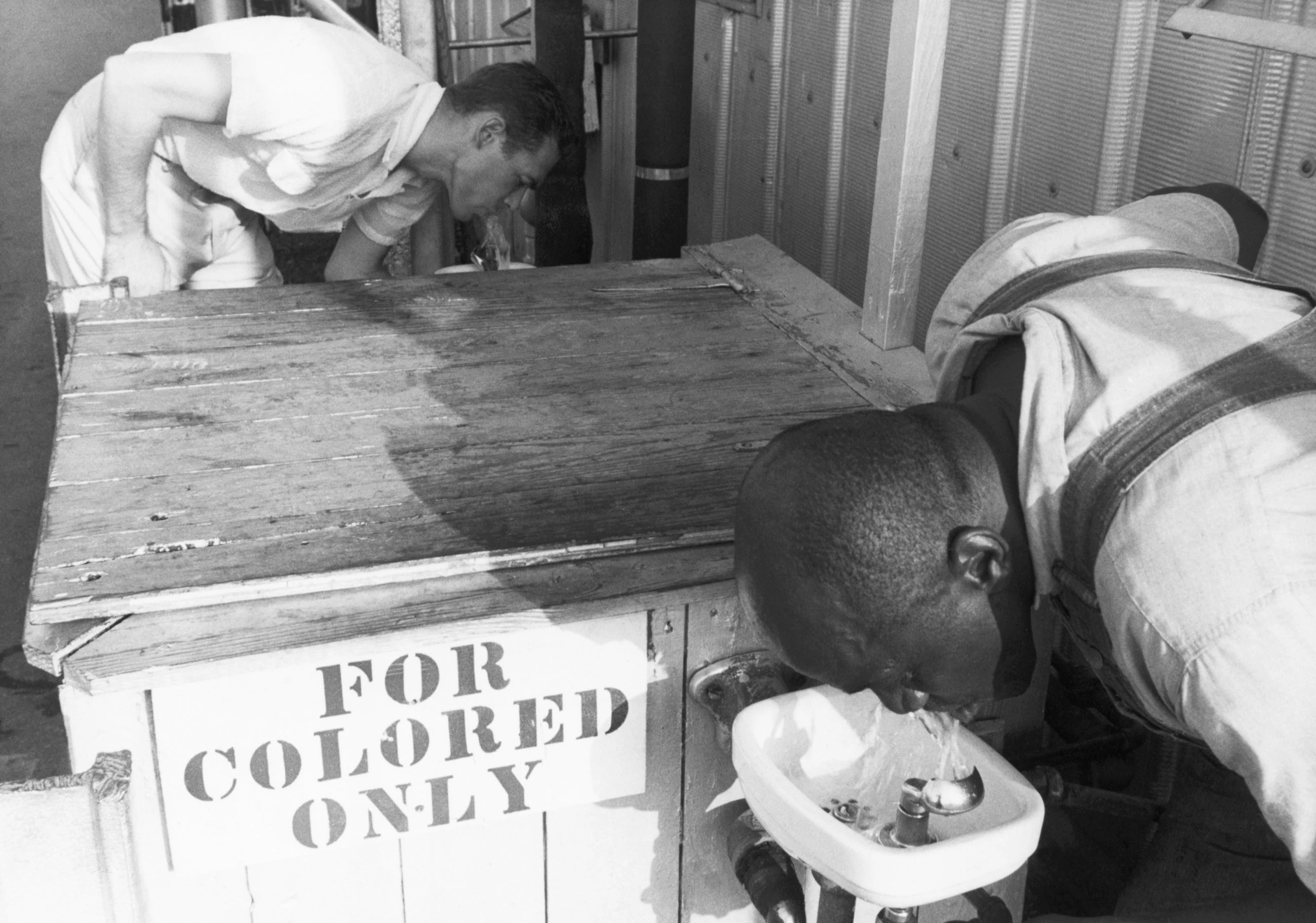 A white man and a Black man drink from separate fountains at the same moment. The nearer fountain is marked “For Colored Only.” The archive dates the image to about 1960; its photographer and precise location are unknown.
