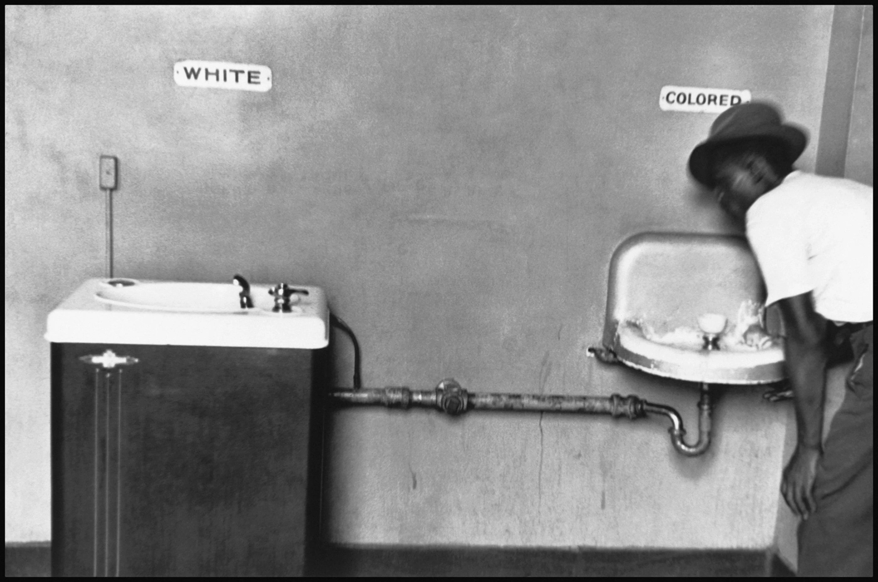 A Black man drinks from a worn basin marked “Colored.” Beside it stands a larger fountain marked “White”; a shared drainpipe is visible beneath them. Photograph by Elliott Erwitt, 1950.