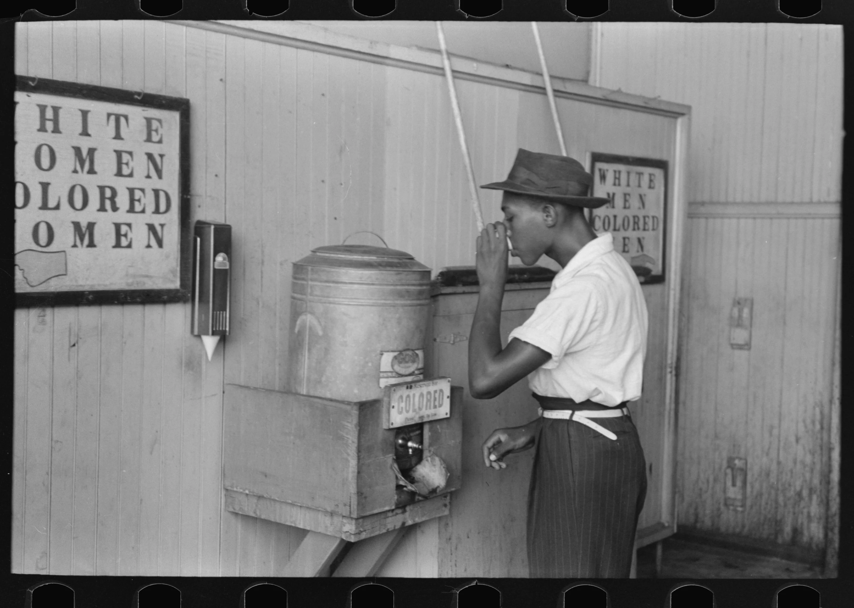 A man drinks from a paper cup beside a cooler marked “Colored.” Signs for white and Black women and men hang on the wall. The archival record does not name him. Photograph by Russell Lee.