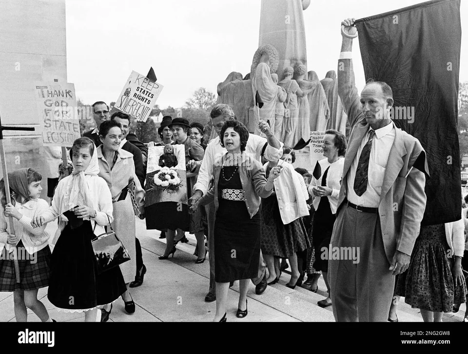 The procession passes a monument near the Capitol. The coffin remains visible behind a woman holding a flag.